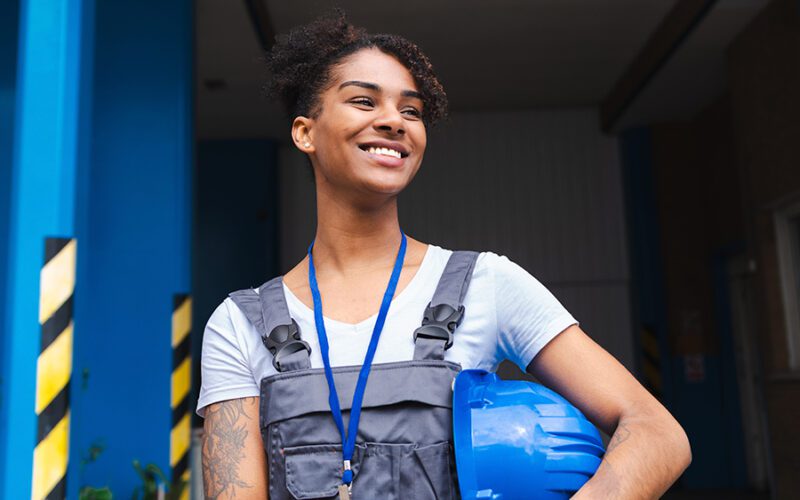 A woman wearing overalls and a lanyard stands holding a blue hard hat, smiling, with a building entrance in the background.