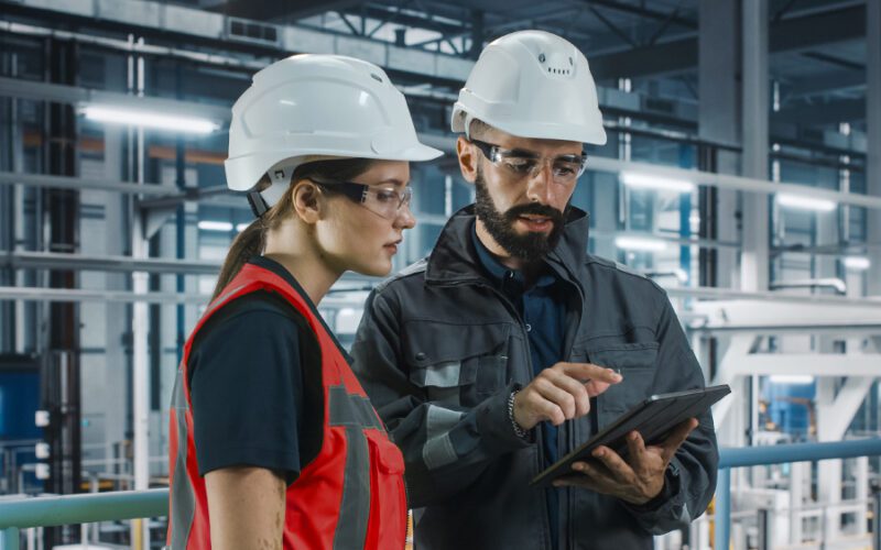 Two workers wearing hard hats and safety glasses stand indoors at an industrial facility, with one pointing at a tablet while discussing information.