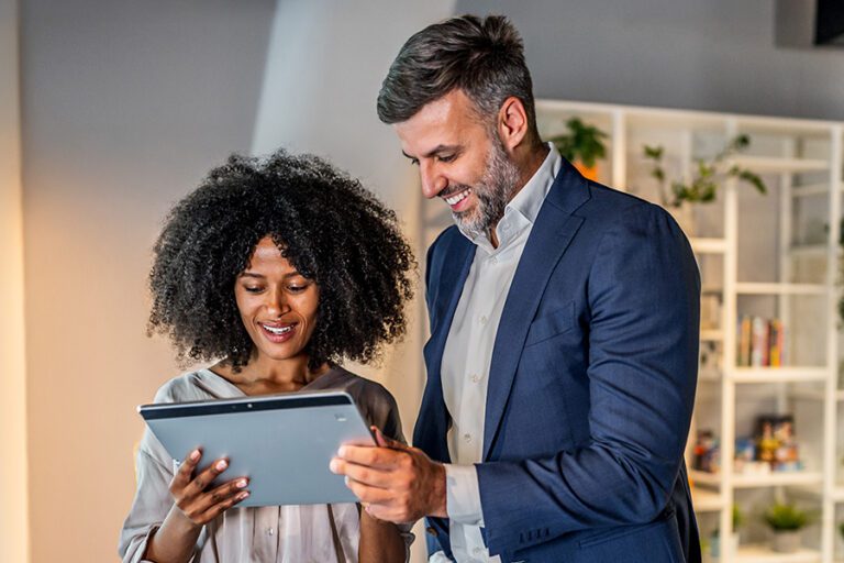 Two professionals, a woman and a man, stand together in an office while reviewing information on a digital tablet.