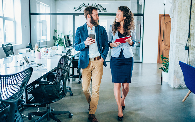 Two business professionals walk through a modern office, holding a coffee cup and a tablet, engaged in conversation.