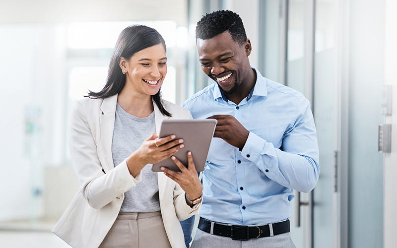 Two people in business attire stand in a hallway, smiling and looking at a tablet device together.