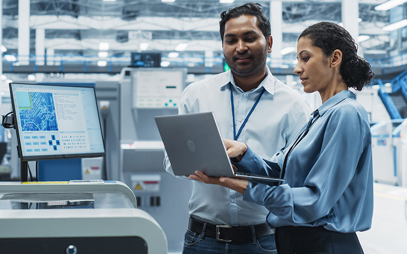 Two people in professional attire review data on a laptop in a modern industrial facility, with machinery and computer monitors in the background.