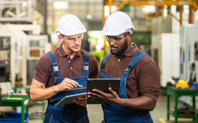 Two factory workers in safety helmets and overalls review information on a tablet and clipboard in an industrial setting.