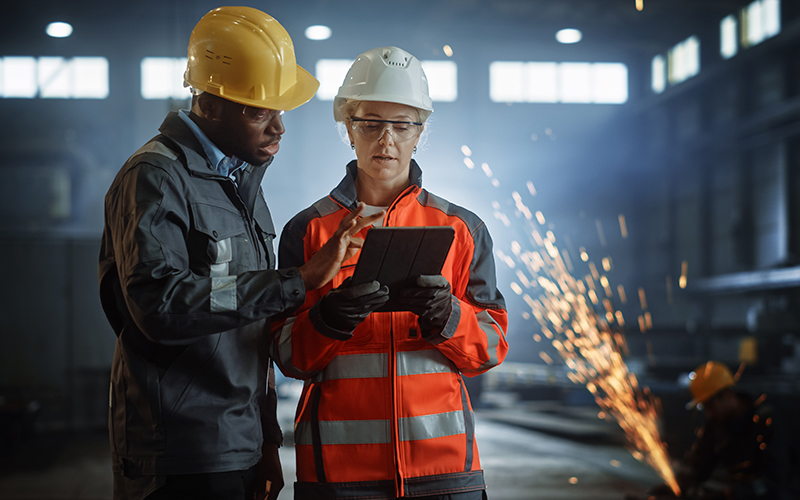 Two industrial workers in safety gear stand in a factory, looking at a tablet together while sparks fly in the background.