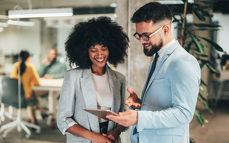 Two professionally dressed people stand indoors, looking at a tablet and smiling, with an office environment and colleagues in the background.