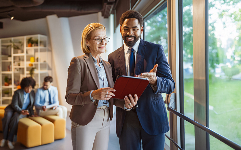 Two business professionals stand by a window discussing information on a tablet, while two colleagues talk in the background in a modern office setting.