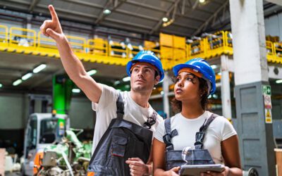 Two factory workers wearing blue hard hats and work overalls stand indoors; one points upward while the other, focused on workforce management tasks, holds a tablet and looks in the same direction.