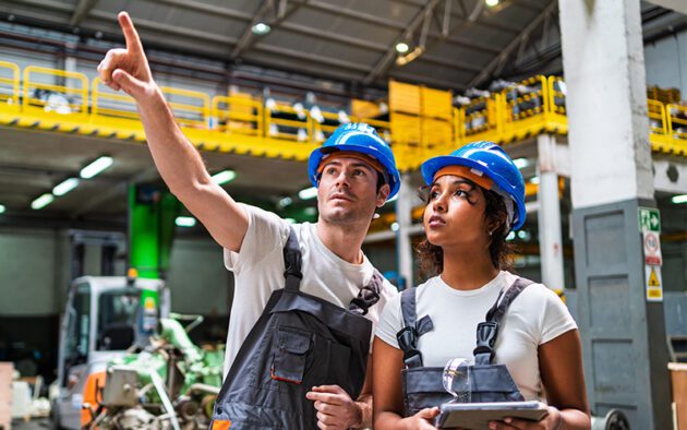 Two factory workers wearing blue hard hats and work overalls stand indoors; one points upward while the other, focused on workforce management tasks, holds a tablet and looks in the same direction.