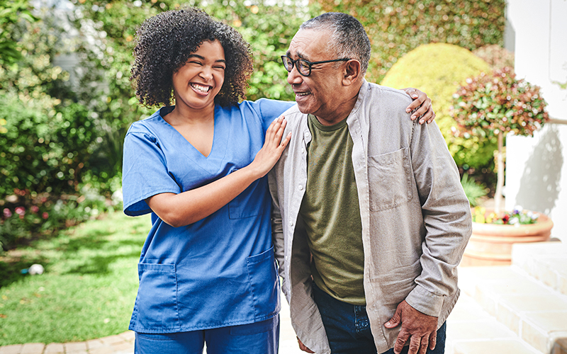 A nurse in blue scrubs stands outside with an older man, smiling and supporting him as they walk together in a garden setting.