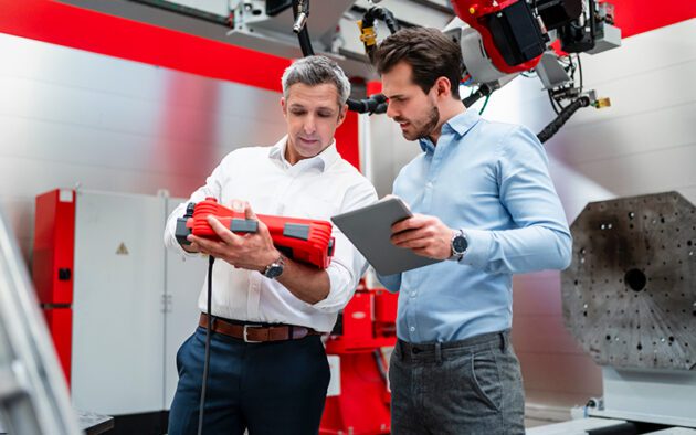 Two men in business attire examine a red industrial device while one holds a tablet in a modern manufacturing or laboratory setting.