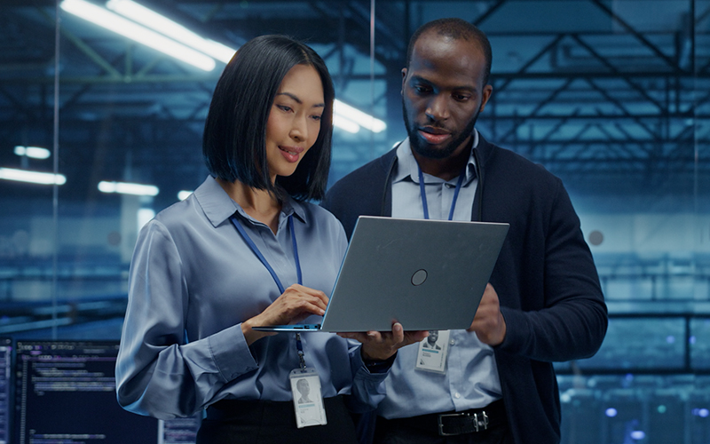 Two professionals wearing ID badges stand together in an office, looking at a laptop screen and discussing workforce management strategies.