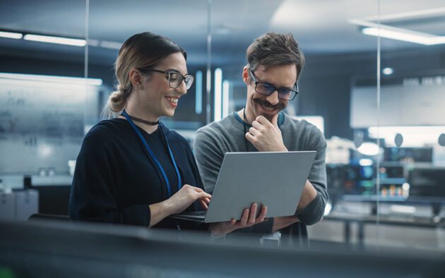 Two people in an office setting stand together, looking at and discussing something on a laptop, surrounded by modern technology and glass walls.