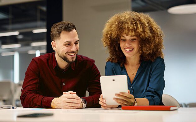 Two people sitting at a table in an office, smiling and looking at a tablet device together.