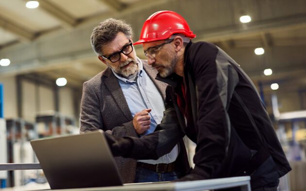 Two men in an industrial setting look at a laptop; one wears a suit and glasses, the other wears a red hard hat and black workwear.