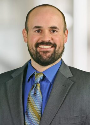 A man with a beard and short hair, wearing a gray suit, blue shirt, and striped tie, smiles at the camera against a blurred light background.