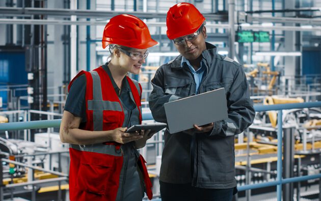 Two workers wearing red helmets and safety gear review information on a tablet and laptop inside an industrial facility with machinery in the background.