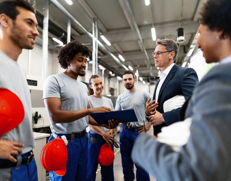 A group of workers in blue uniforms and holding red helmets talks with two people in business attire inside an industrial facility.