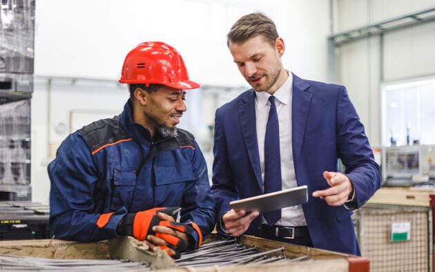 A man in a suit shows a tablet to a worker wearing a red hard hat and gloves in an industrial setting.