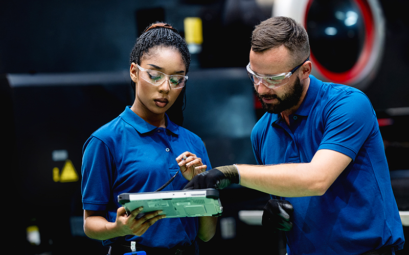 Two factory workers in blue uniforms and safety goggles review information on a tablet while standing in an industrial setting.