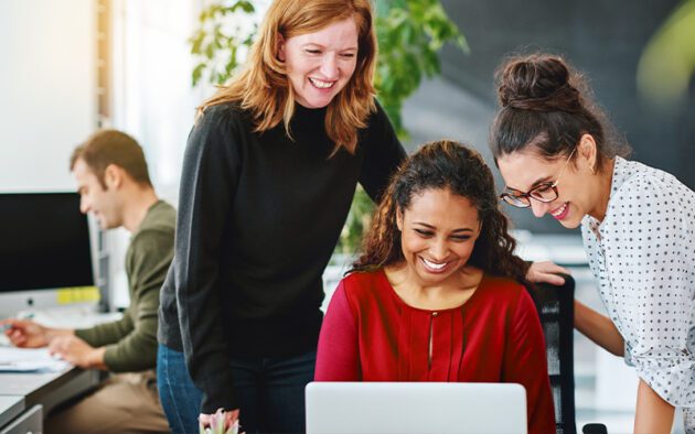 Three women smile and look at a laptop screen together in an office, while a man works at a desk in the background.