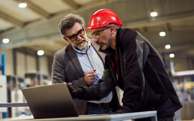 Two men in a factory setting, one in a suit and glasses, the other in a red hard hat, look at a laptop screen on a workbench.