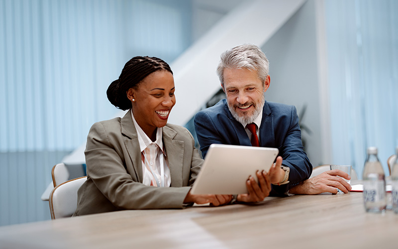 Two business professionals sit at a table, smiling and looking at a tablet together during a meeting in a modern office setting.