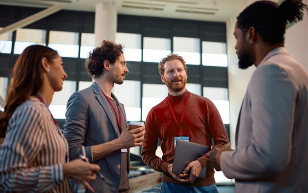 Four people wearing conference badges stand in a modern office space, engaged in conversation. One holds a coffee cup, another holds a laptop.