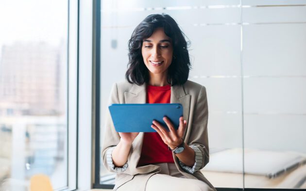 A woman in a beige blazer and red shirt sits by a window, smiling while using a blue tablet.