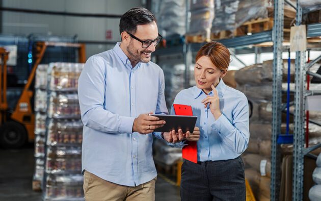 Two people in business attire review information on a tablet while standing in a warehouse with shelves and boxes in the background.