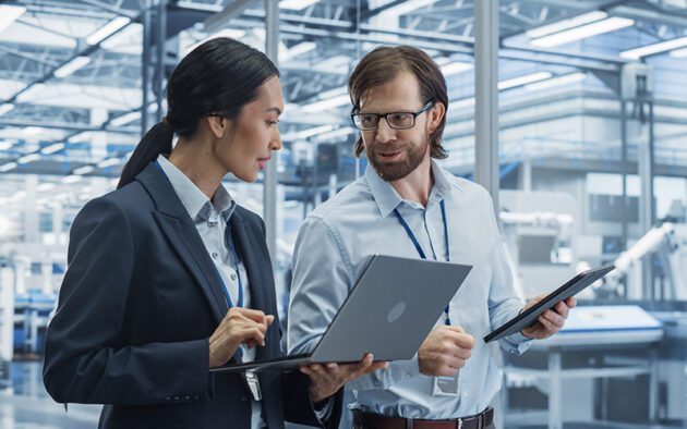 Two professionals in an industrial setting discuss work while holding a laptop and a tablet, with machinery and large windows visible in the background.