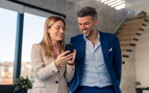 Two business professionals stand indoors, smiling while looking at a smartphone together. Modern office setting in the background with large windows and a staircase.