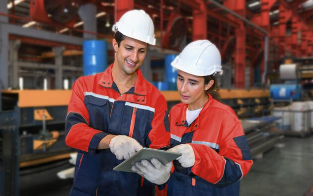 Two workers in safety gear and helmets stand in a factory, looking at a tablet together. Industrial equipment and machinery are visible in the background.
