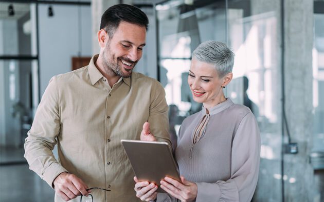 Two colleagues standing in an office, smiling and looking at a tablet together.