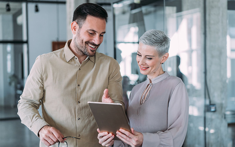 Two colleagues standing in an office, smiling and looking at a tablet together.