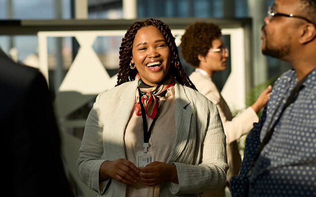 A woman wearing a light blazer and lanyard smiles while interacting with others at a professional indoor event.
