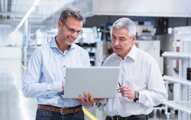 Two men in business attire stand in a modern industrial setting, looking at a laptop and discussing its contents.