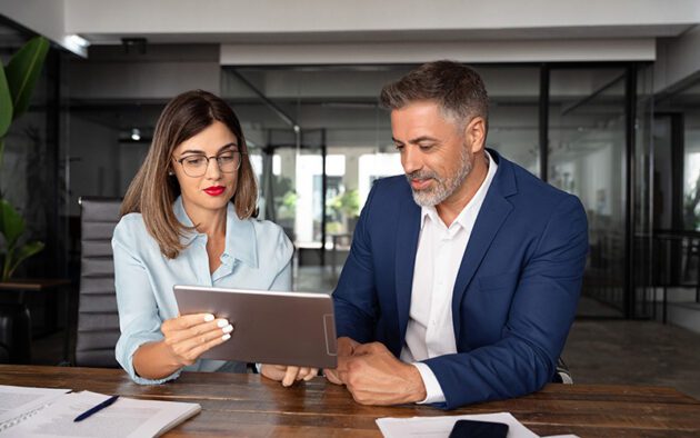 Two people sitting at a desk in an office, reviewing information on a tablet together.
