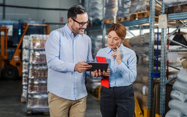 Two people in business attire review information on a tablet in a warehouse, surrounded by shelves stocked with goods.
