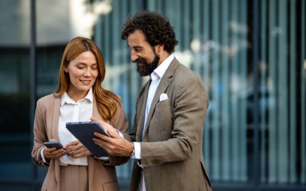 Two business professionals standing outdoors, one holding a tablet and the other holding a smartphone, both dressed in suits and engaged in a discussion.