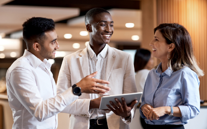Three professionally dressed people stand together indoors, having a discussion while one of them holds a digital tablet.