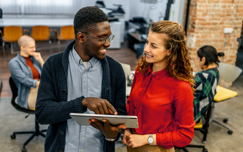 Two colleagues stand and smile while discussing something on a tablet in a modern office setting, with other people working in the background.