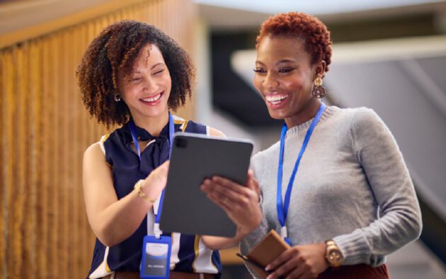 Two women wearing lanyards stand together, smiling and looking at a tablet device indoors.