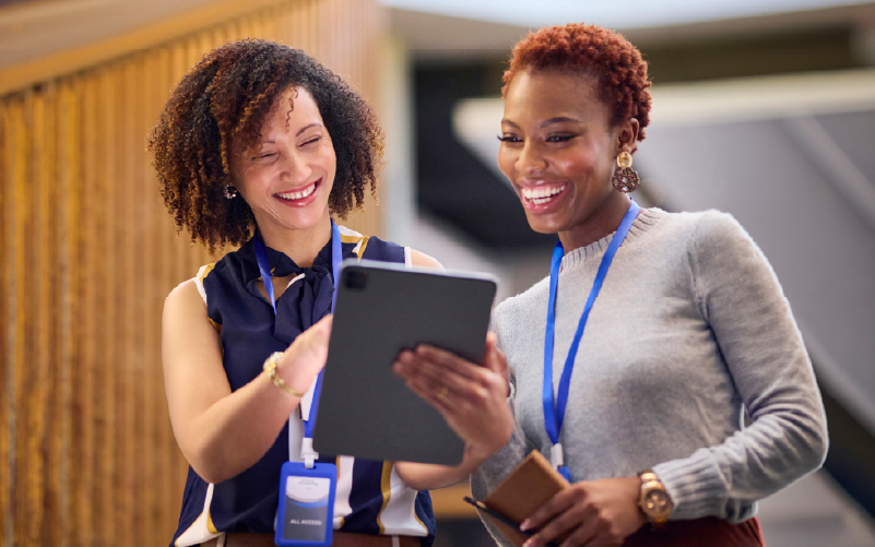 Two women wearing lanyards stand together, smiling and looking at a tablet device indoors.