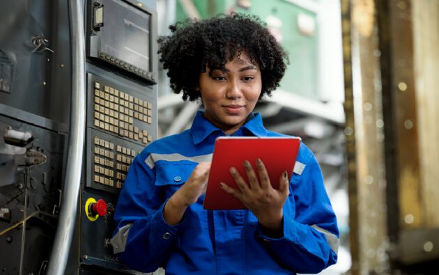 A person in a blue work uniform uses a red tablet while standing next to industrial machinery.