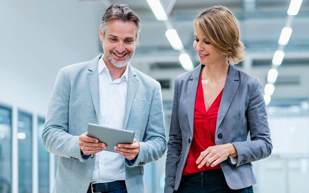 Two business professionals, a man and a woman, stand indoors looking at a tablet device together. Both are dressed in business attire and appear to be discussing something.