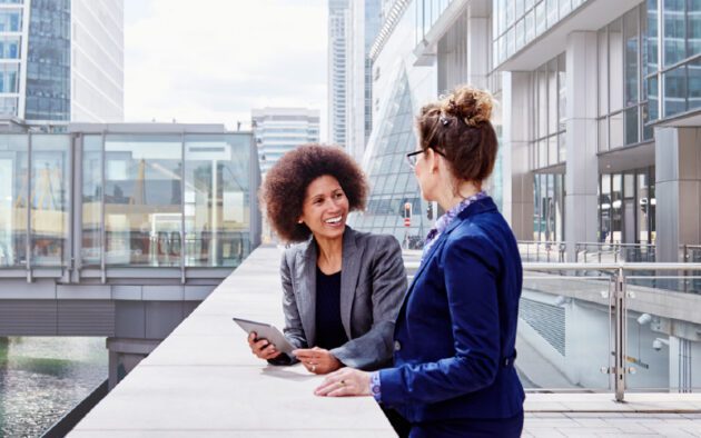 Two women in business attire have a conversation outdoors in an urban setting, with modern office buildings in the background. One woman holds a tablet.