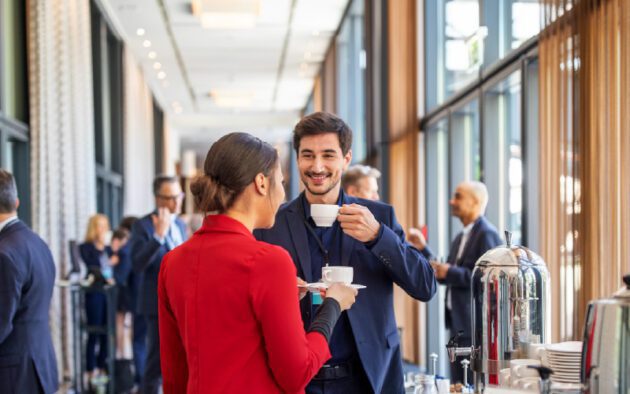 People in business attire conversing and drinking coffee at a networking event in a bright, modern conference space.