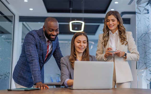 Three business professionals stand around a desk, looking at a laptop screen and smiling in a modern office setting.