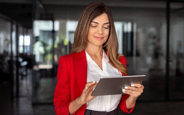 A woman in a red blazer stands indoors, smiling while using a tablet—showcasing the power of technology in today’s global workforce.