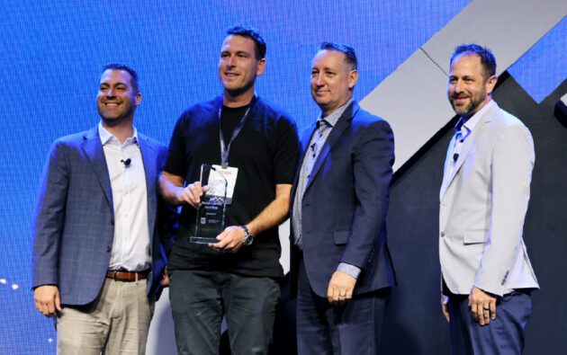 Four men stand on stage, one holding a glass award, all dressed in business attire. Against the blue-lit background, they celebrate achievements in workforce management.